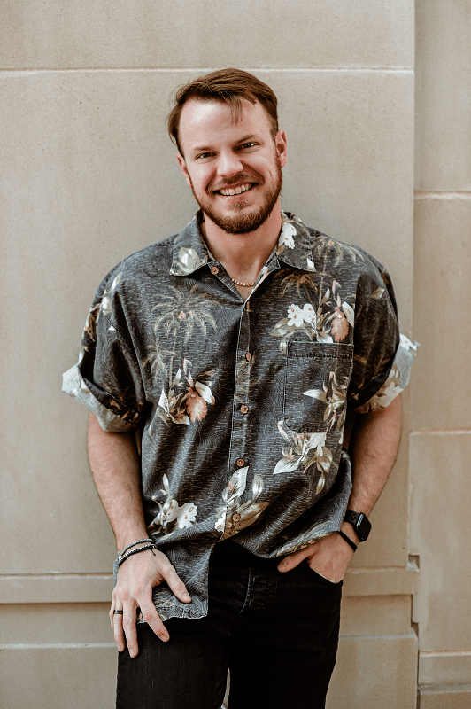 A white man in a floral, short-sleeve button up shirt leaning against a wall.