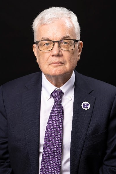 A professional headshot of Dr. Stephen Addison, an old man with white hair wearing a purple tie and a UCA Now pin.