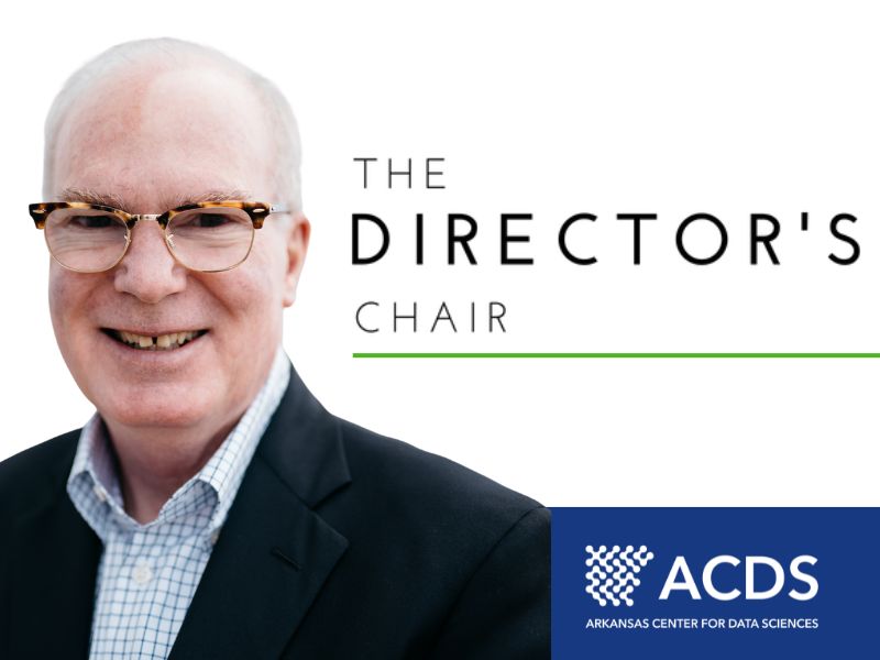 A headshot of an old white man with white hair on the sides of his head wearing glasses, a black suit jacket, and a blue and white checkered shirt on the Director's Chair announcement.