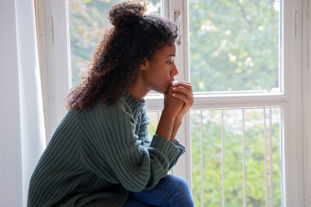 A black woman wearing a green sweater sitting by a window with her hands folded, propped against her chin.