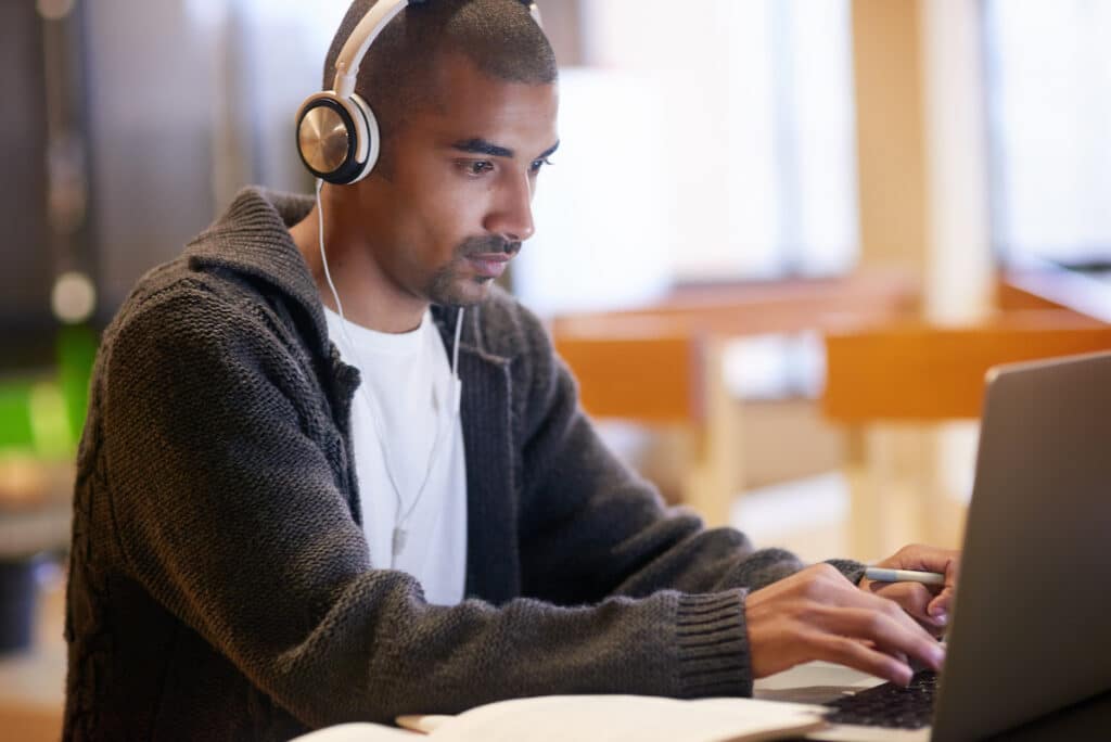 A young man with a shaved head sits at his laptop wearing headphones and a gray sweater.