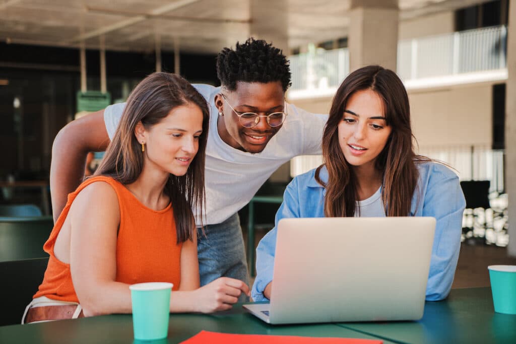 A group of 3 multiracial happy students using a laptop at a green picnic table.