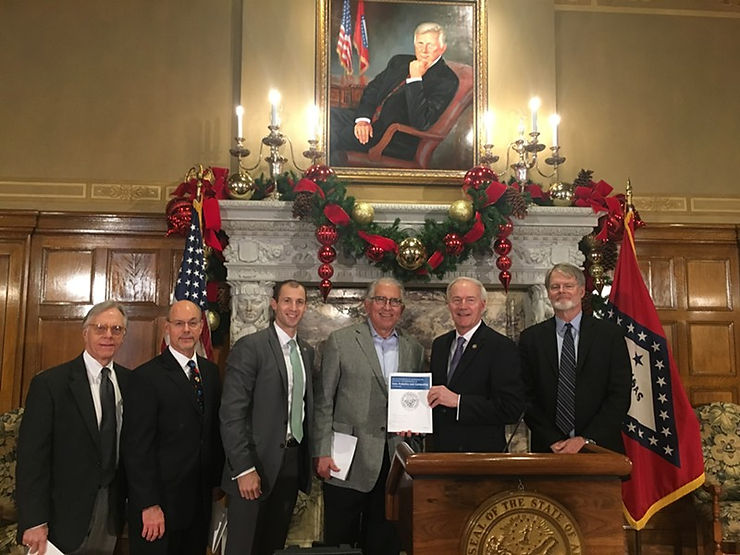 Governor Asa Hutchinson and other leaders stand in front of a large marble fireplace with a picture of bill clinton hanging above it.