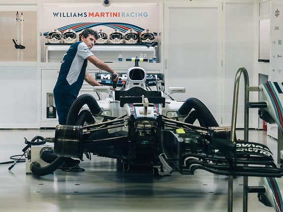 A man working on a large machine in a room. Behind his head a sign says "William Martini Racing" and there are multiple helmets sitting on the shelf below.