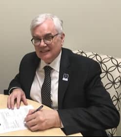 An old man with white hair sitting in a fabric covered chair, wearing a suit and signing a white piece of paper.