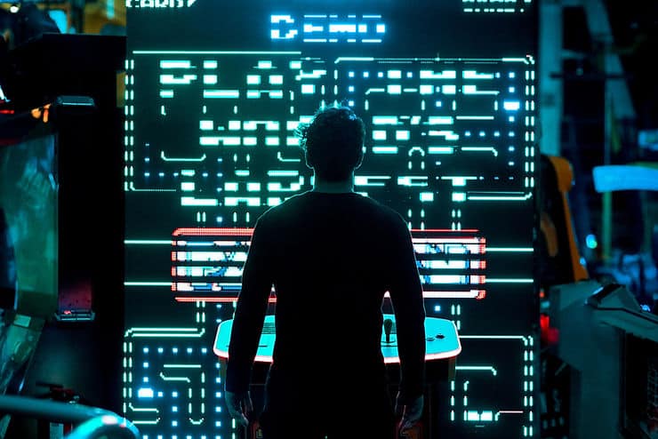 A man standing in front of a large pacman game. The room is dark but the screen lights up the silhouette of the man with neon blue light.