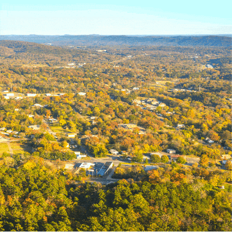A motion graphic of an arial view of neighborhoods interspersed with copses of trees changing colors. Small black location pins zoom in and off the screen with a message that says "Empowering You. Transforming Arkansas."