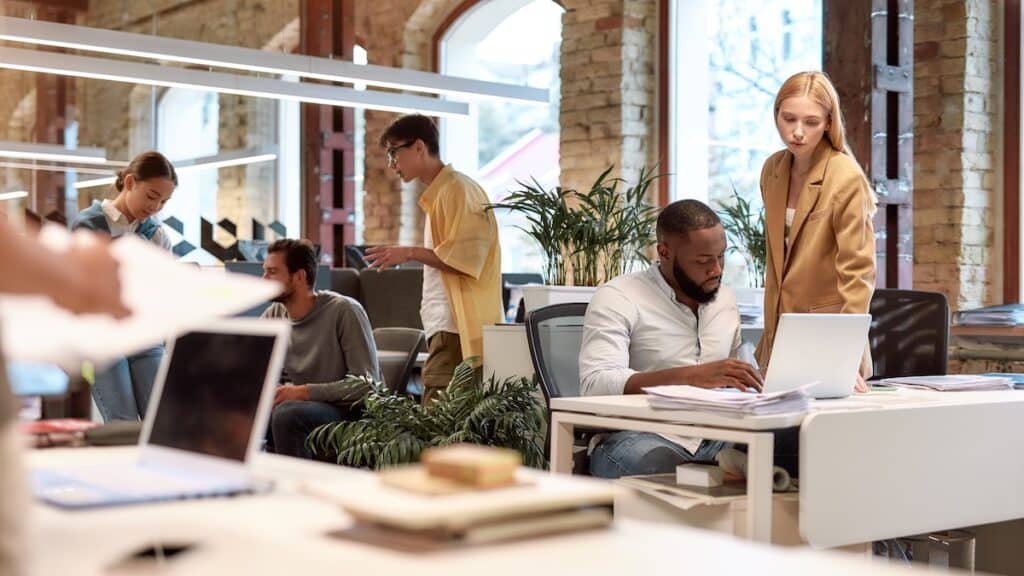 A brightly lit office space with large windows, plants, and spacious desks. Employees sit and stand at the desks, looking at their work and collaborating.