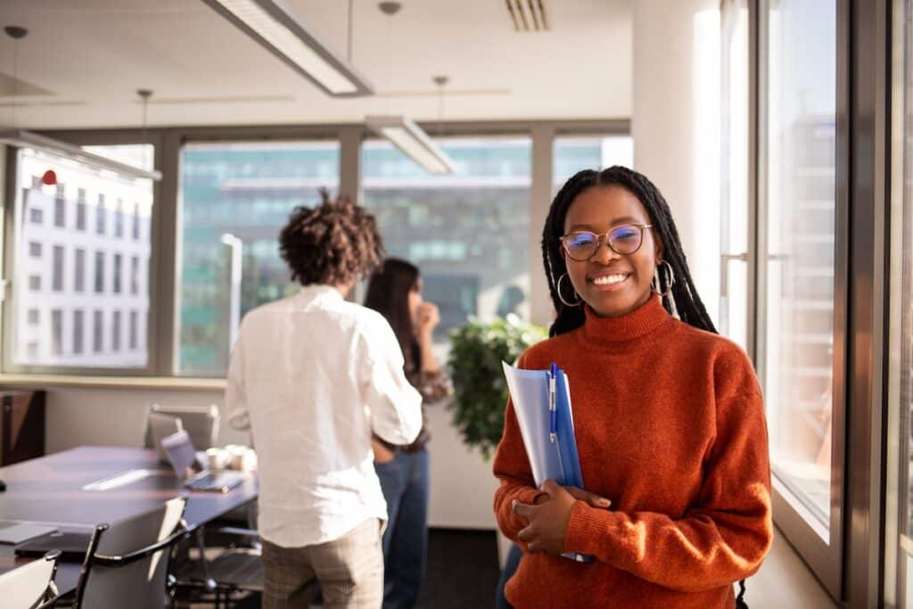 A black girl in an office in the foreground is looking at camera. She is wearing a burnt orange sweater and is holding a blue folder..